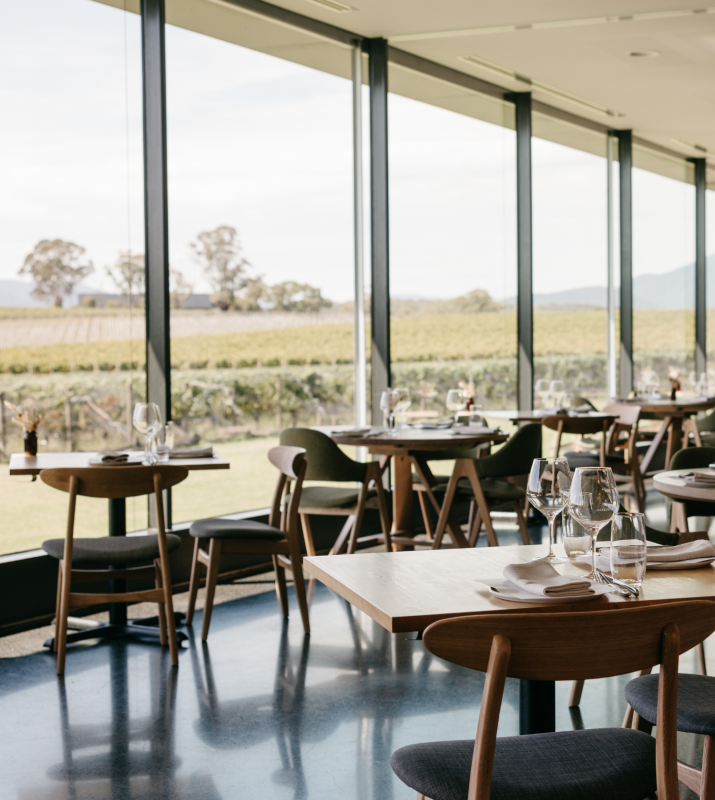The dining room of Oakridge Restaurant in the Yarra Valley. The scene shows polished wooden tables, set for restaurant dining, and floor to ceiling windows which take in the expansive vineyard views.